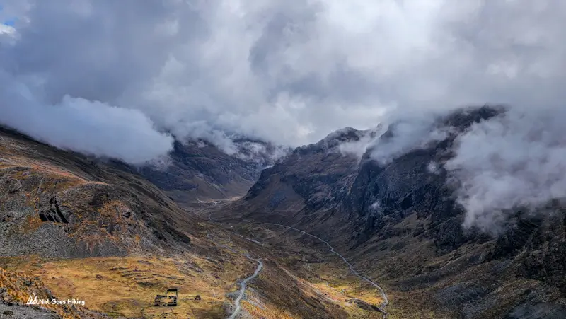 Winding road and stone ruin in a cloud-filled Andean valley with dark ridges on the El Choro Trek