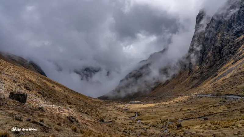 Wide Andean valley with puna grass, a small stream and dark cliffs wrapped in cloud on the El Choro Trek