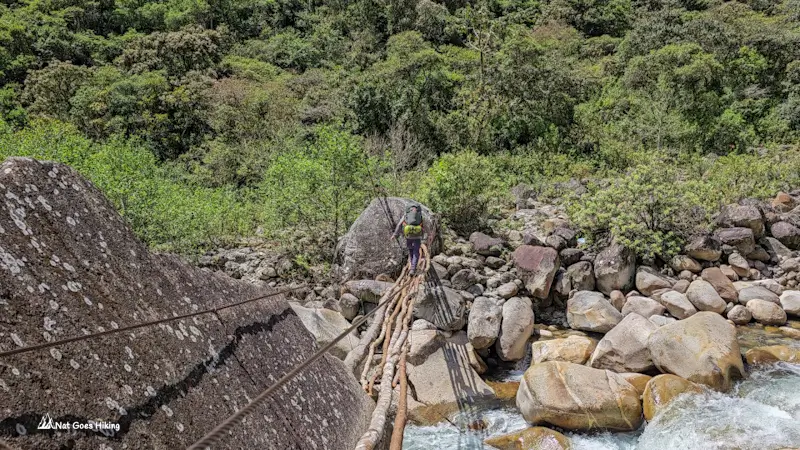 Hiker with a backpack crossing a narrow pole bridge over the Chucura River between boulders on the El Choro Trek