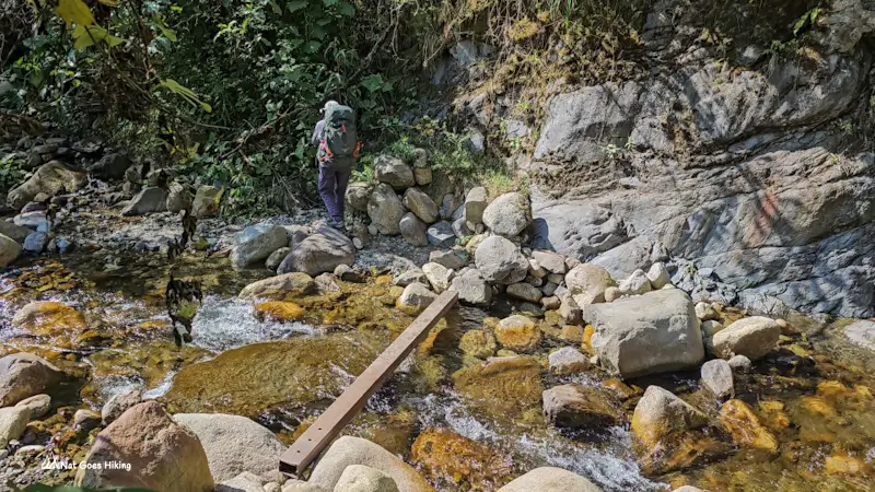Hiker with a backpack beyond a single metal plank laid across a shallow rocky stream on the El Choro Trek